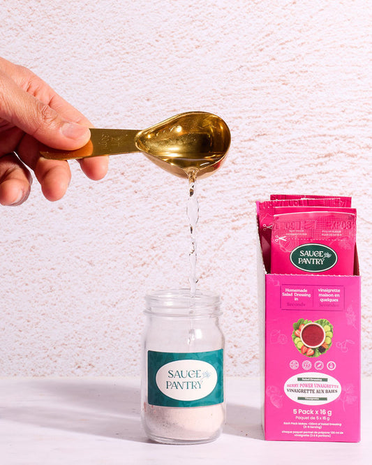 Hand pouring a spoonful of a white powder into a small bottle with a pink packet labeled 'Nature's Plus' on a light background.