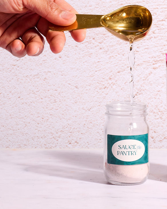 Hand pouring a liquid from a gold spoon into a glass jar labeled 'Sauce Pantry' on a textured white background.
