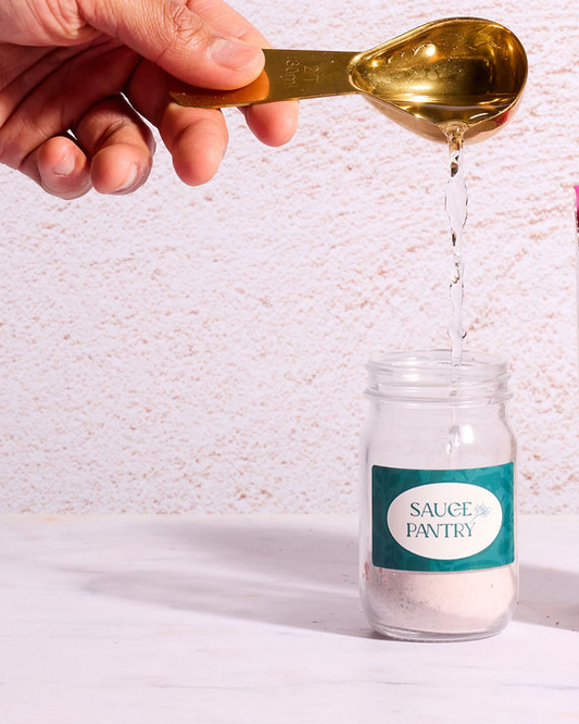 Hand pouring a liquid from a gold spoon into a glass jar labeled 'Sauce Pantry' on a textured white background.