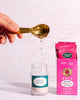 Hand pouring a spoonful of a white powder into a small bottle with a pink packet labeled 'Nature's Plus' on a light background.